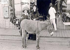 A woman and a man, wearing a hat and a Mexican shawl, stand behind a donkey and in front of a souvenir shop.